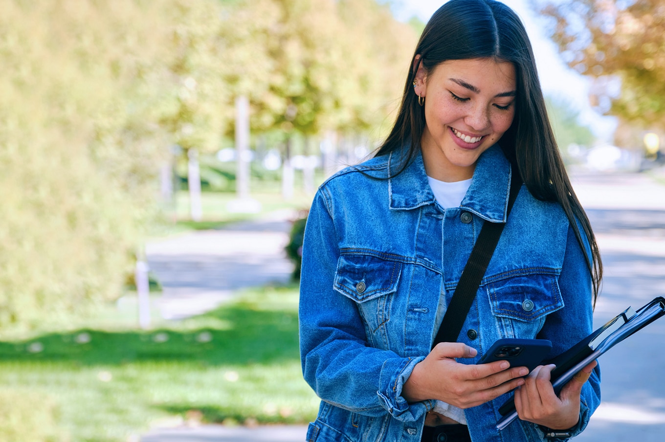 Smiling woman in a denim jacket looking at her phone while holding a notebook outdoors.