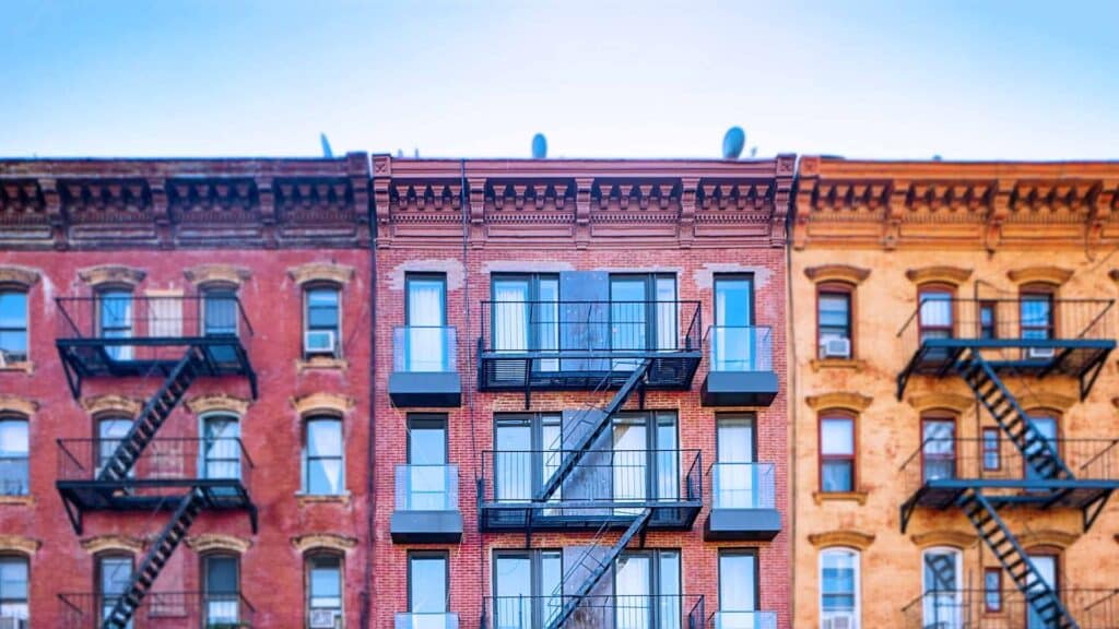 Row of colorful brick apartment buildings with black metal fire escapes and balconies against a clear blue sky.
